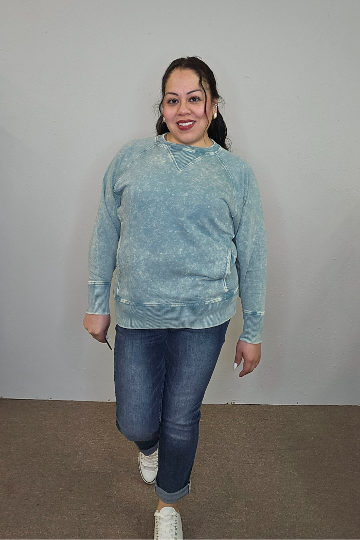 A woman stands indoors against a plain light wall, smiling. She wears the Acid Wash Terry Pullover in light blue with blue jeans and white sneakers, her hair pulled back and one foot stepping forward.