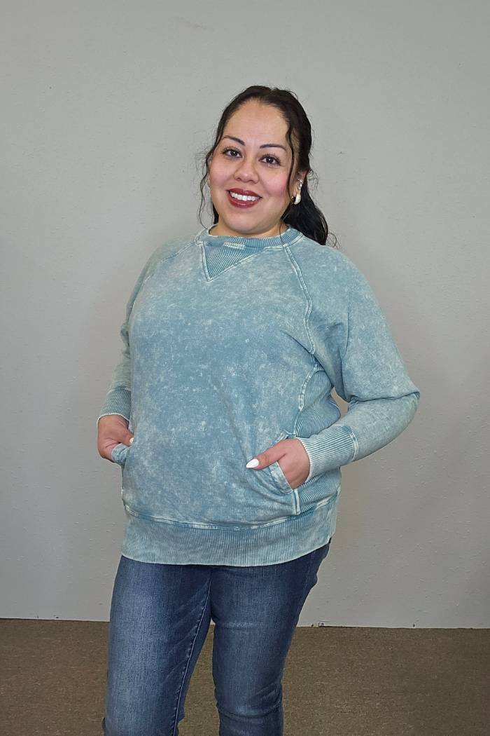 A woman smiles against a light gray wall, wearing an oversized Acid Wash Terry Pullover in light blue with her hands in the pockets and blue jeans. Her dark hair is tied back, and she wears light makeup.