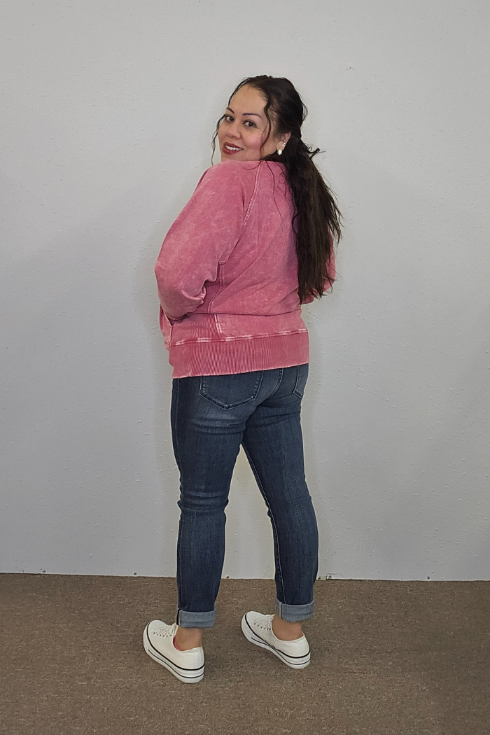 A woman with long dark hair, smiling and turned slightly away from the camera, wears an oversized pink Acid Wash Terry Pullover with dark blue jeans and white sneakers. She stands on a brown carpet against a plain light gray wall.