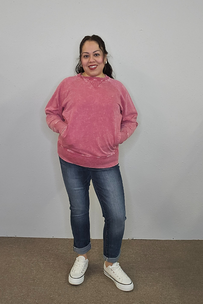 A woman stands against a plain light gray wall, smiling. She wears an Acid Wash Terry Pullover, blue cuffed jeans, and white sneakers. Her dark hair is pulled back and her hands are in her sweatshirt pockets.