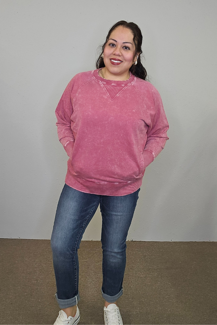 A woman smiles with her hands in the pockets of her oversized Acid Wash Terry Pullover, paired with blue rolled-up jeans and white sneakers, standing against a plain light gray wall.