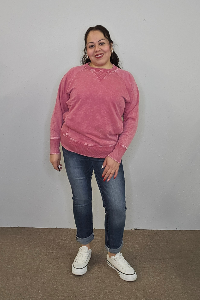 A woman with long dark hair, wearing the Acid Wash Terry Pullover, blue jeans, and white sneakers, stands smiling against a plain light gray wall and brown carpeted floor.
