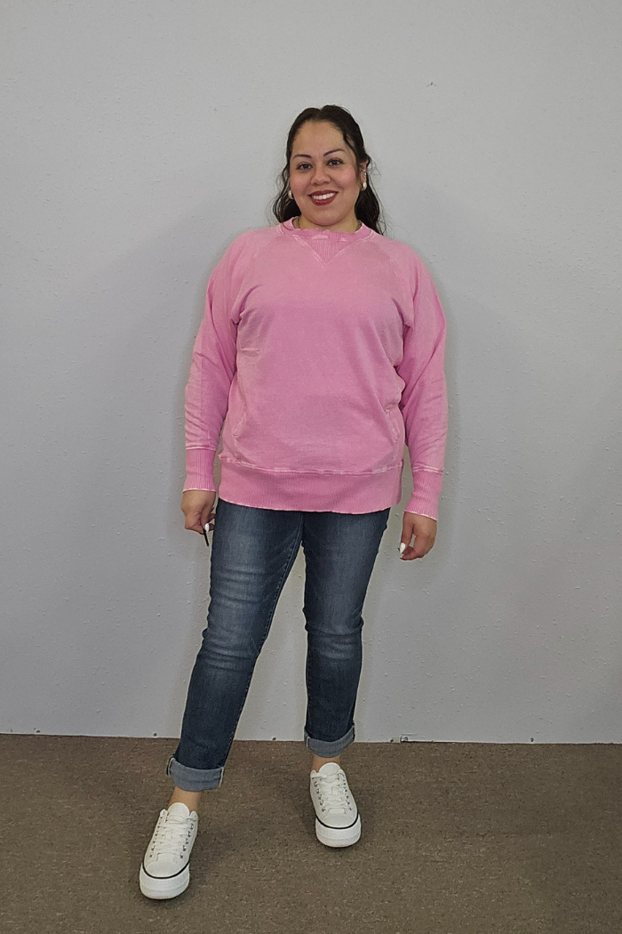 A woman stands indoors against a light gray wall, smiling in an oversized pink Acid Wash Terry Pullover, rolled-up blue jeans, and white sneakers. She poses with one leg slightly forward.