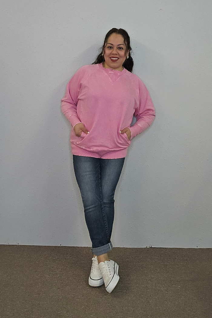A woman stands against a plain light gray wall, smiling. She wears an Acid Wash Terry Pullover with front pockets, blue rolled-up jeans, and white sneakers. Her hands are in her pullover pockets and her hair is pulled back.