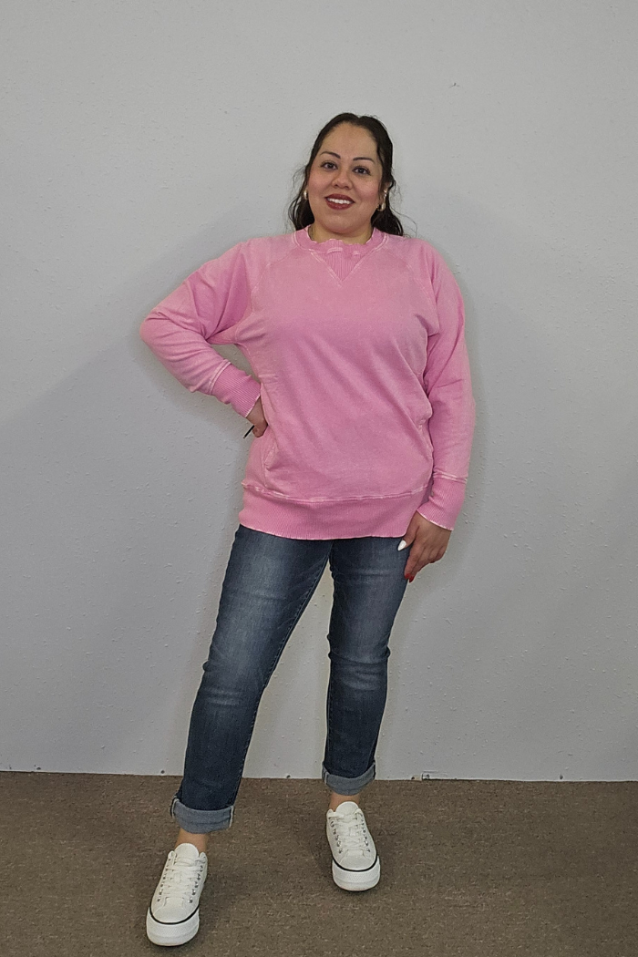 A woman stands against a light gray wall, smiling in an Acid Wash Terry Pullover, blue rolled-up jeans, and white sneakers. Her dark hair is tied back and one hand rests on her hip.