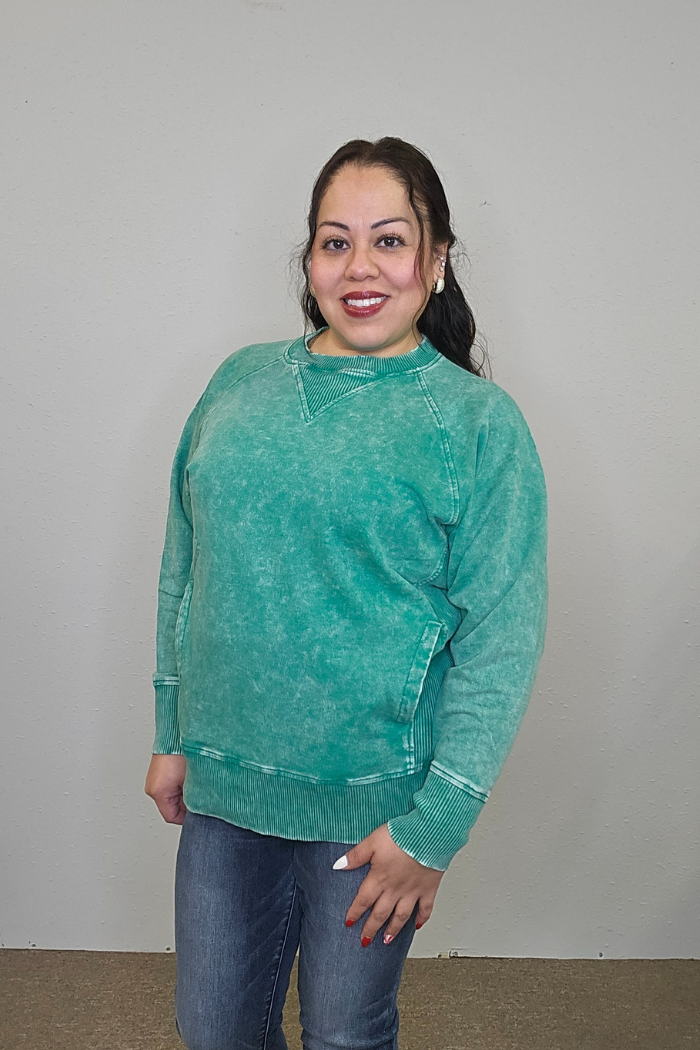 A woman with long dark hair smiles in front of a plain light wall, wearing the Acid Wash Terry Pullover in green, blue jeans, and showing off her white nails.