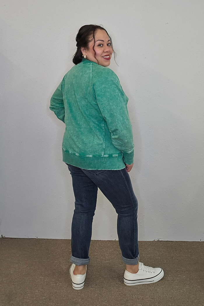 A woman with dark hair in a bun, wearing an oversized Acid Wash Terry Pullover, dark cuffed jeans, and white sneakers smiles and looks over her shoulder against a plain light-colored wall.