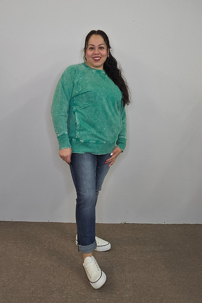 A woman with long dark hair, in an oversized Acid Wash Terry Pullover, blue jeans, and white sneakers stands smiling against a plain light-colored wall.