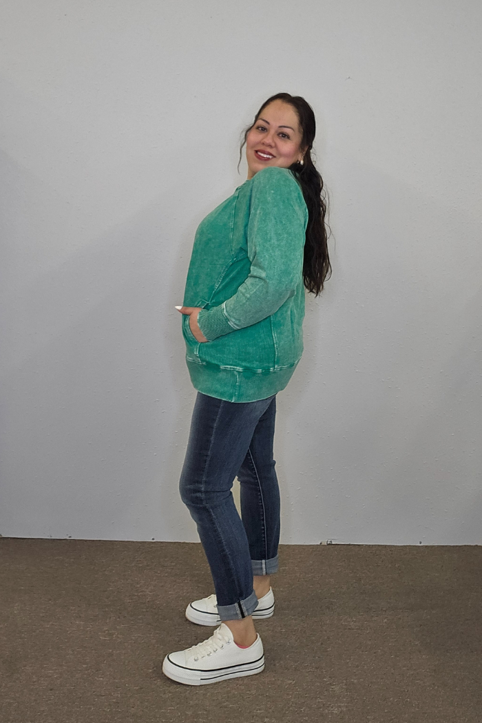 A woman stands sideways, smiling with hands in the pockets of her Acid Wash Terry Pullover, paired with rolled-up blue jeans and white sneakers. Her long dark hair is down, and she poses against a plain light gray wall.