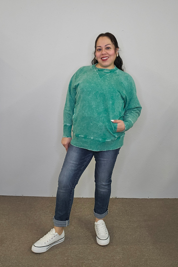 A smiling woman poses in front of a plain light-colored wall, wearing the Acid Wash Terry Pullover in green, paired with rolled-up blue jeans and white sneakers. One hand is in her pocket and her head is tilted slightly.