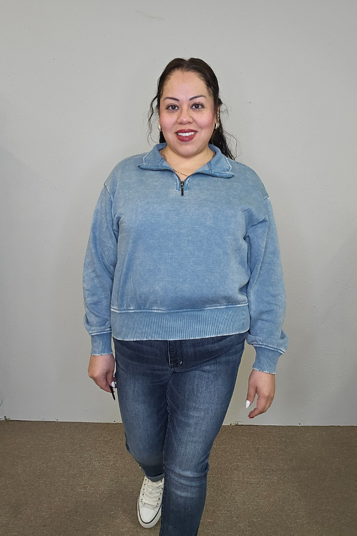 A woman with long dark hair, wearing the light blue Acid Wash Half Zip pullover with blue jeans and white sneakers, smiles in front of a plain light-colored wall.