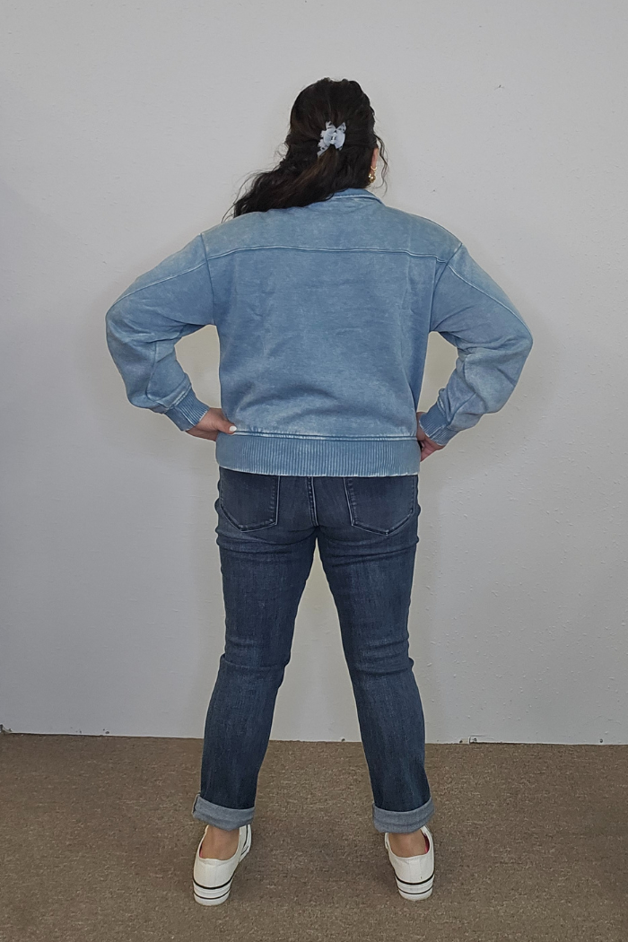 A person with long dark hair in a scrunchie stands facing away, hands on hips, wearing the oversized Acid Wash Half Zip pullover, cuffed blue jeans, and white slip-on shoes against a light wall and brown carpet.
