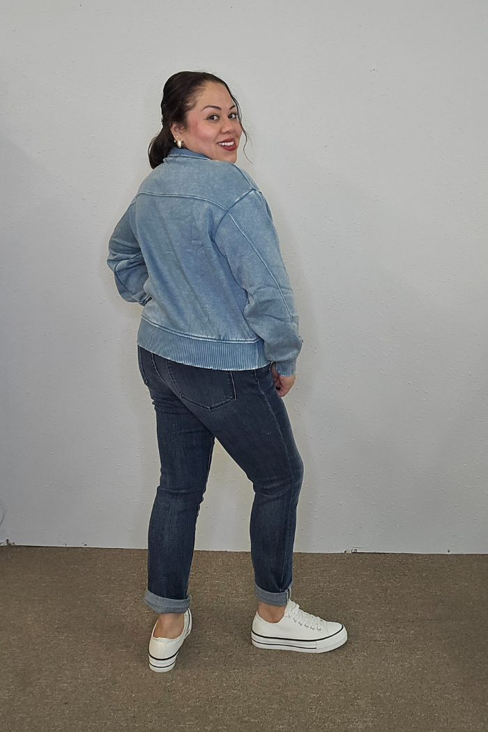 A woman stands indoors, smiling over her shoulder. She wears the Acid Wash Half Zip pullover in light blue, paired with dark rolled-up jeans and white sneakers, set against a plain light-colored wall and carpeted floor.