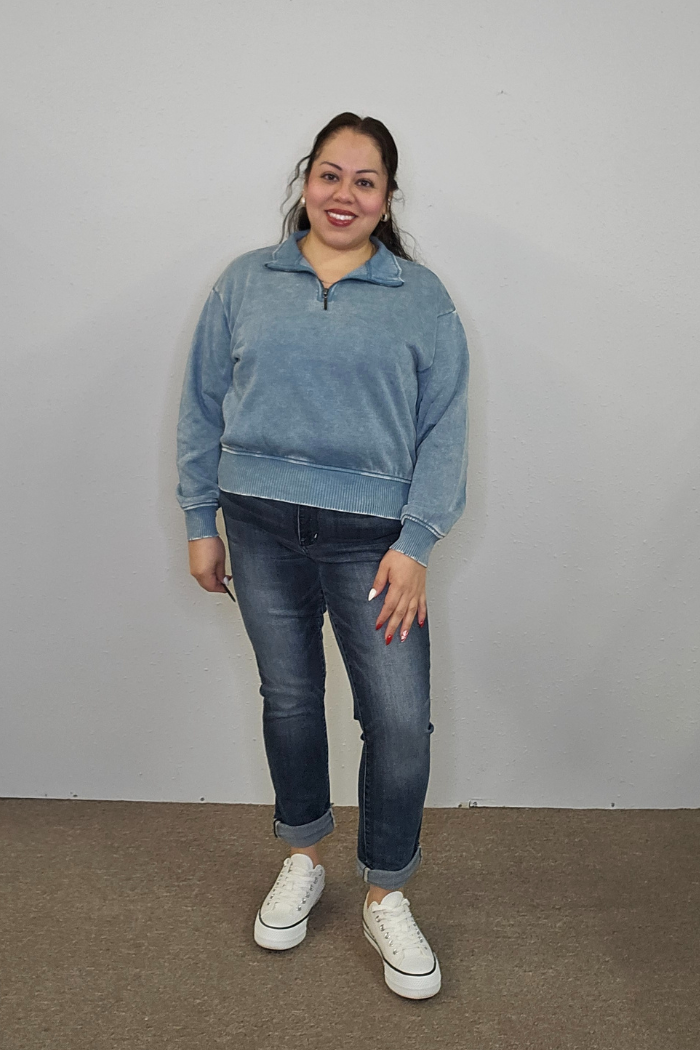 A woman smiles in front of a light gray wall, wearing the Acid Wash Half Zip with cuffed blue jeans and white sneakers. Her dark hair is tied back, and she has red lipstick and painted nails.