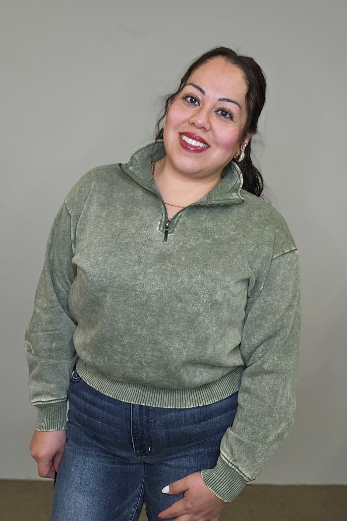 A woman with long dark hair smiles at the camera, wearing the Acid Wash Half Zip pullover in green and blue jeans, standing in front of a plain light gray background.