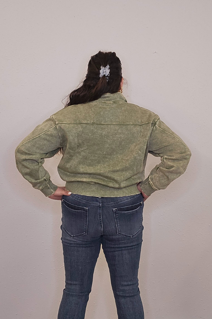 A person with long dark hair in a scrunchie stands hands on hips, facing a plain wall. They’re wearing an oversized, cozy green Acid Wash Half Zip pullover and blue jeans.