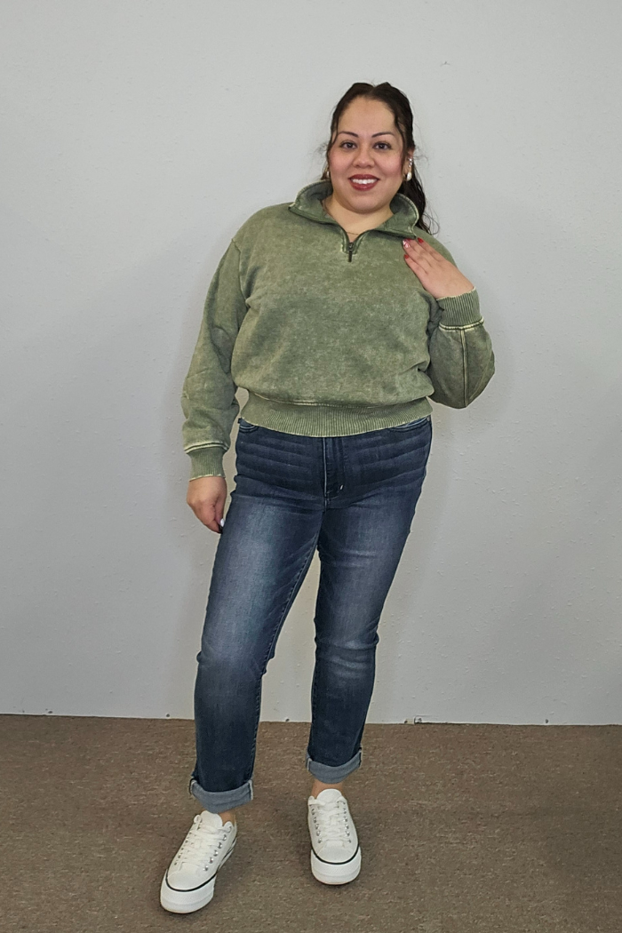 A woman stands against a plain wall, smiling. She wears the Acid Wash Half Zip in green, blue cuffed jeans, and white sneakers. One hand rests on her chest while the other hangs by her side.