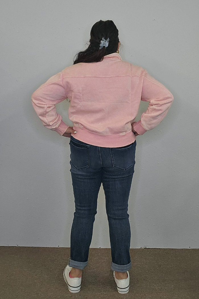A person with long dark hair in a light blue hair clip stands with hands on hips, wearing the Acid Wash Half Zip in pink, dark blue cuffed jeans, and white shoes, facing a plain gray wall.
