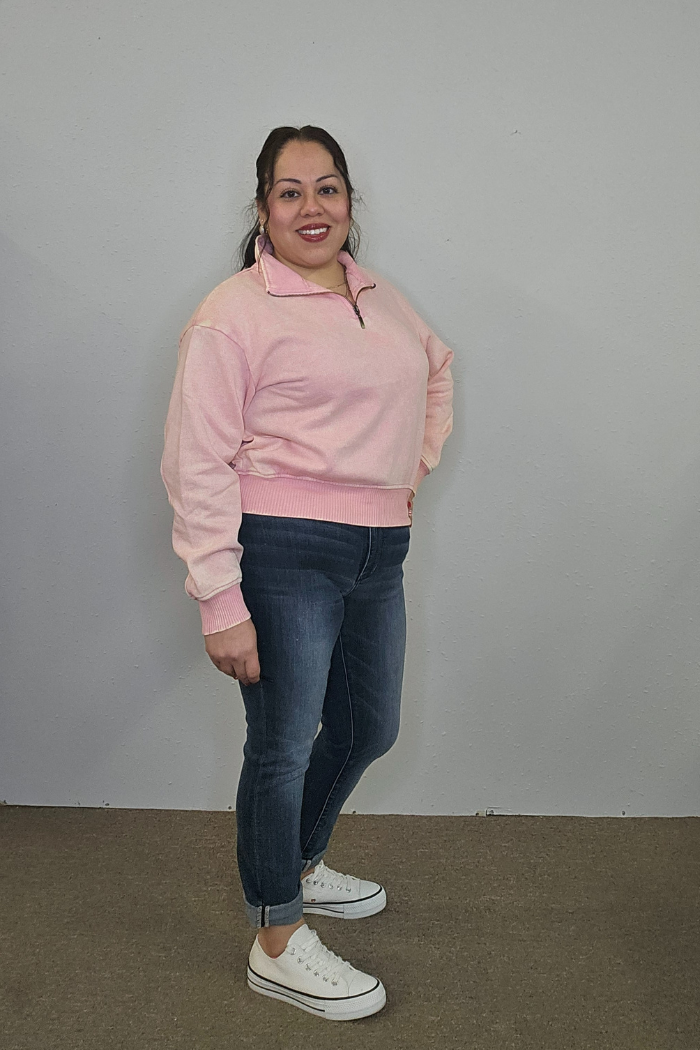 A person stands indoors against a plain wall, smiling. They wear the Acid Wash Half Zip pullover in light pink, blue cuffed jeans, and white platform sneakers. Their hair is pulled back and one hand rests on their hip.