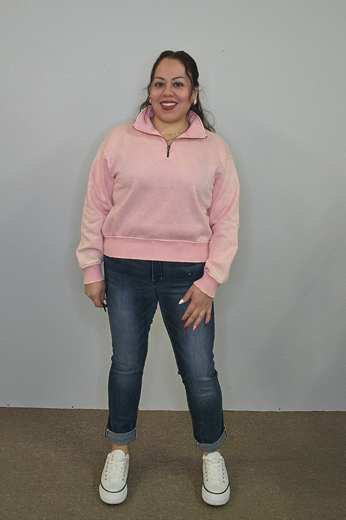 A woman stands against a plain gray wall, smiling. She is wearing the Acid Wash Half Zip pullover in light pink, blue rolled-up jeans, and white sneakers. Her dark hair is tied back, and one hand rests by her side.