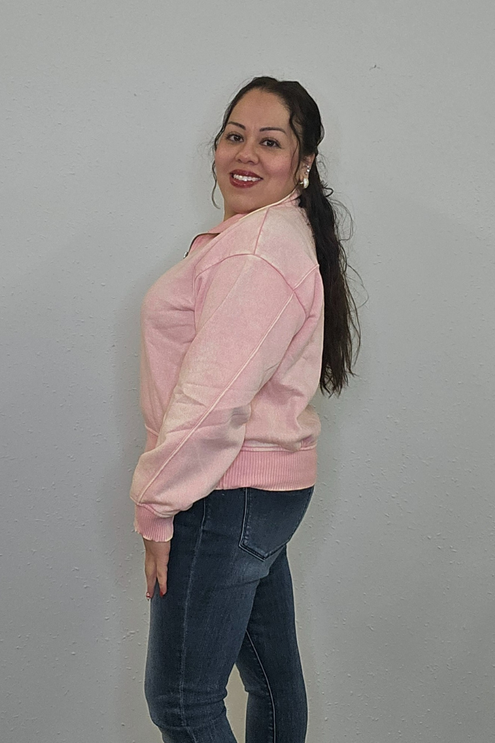 A woman with long dark hair, wearing the Acid Wash Half Zip in light pink and blue jeans, stands sideways and smiles at the camera against a plain light gray wall.