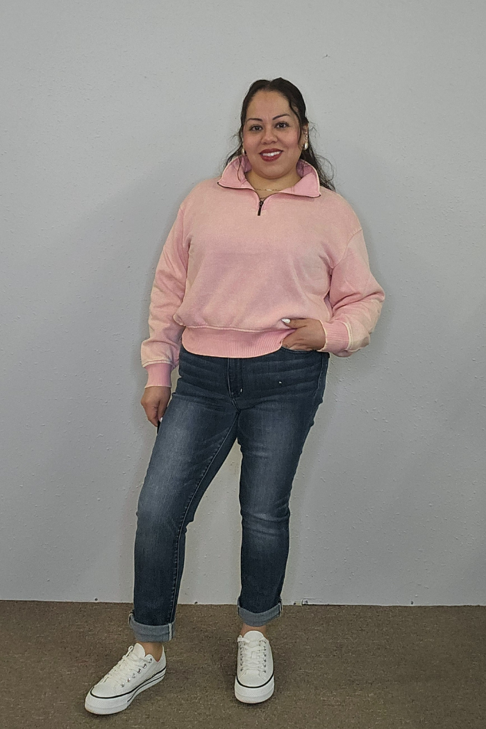 A woman smiles against a gray wall, wearing the Acid Wash Half Zip pullover in light pink, blue cuffed jeans, and white sneakers. One hand is in her pocket while the other rests by her side.