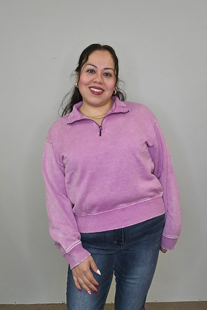 A woman with long brown hair smiles at the camera, wearing the Acid Wash Half Zip pullover in light purple and blue jeans, standing against a plain light gray background.