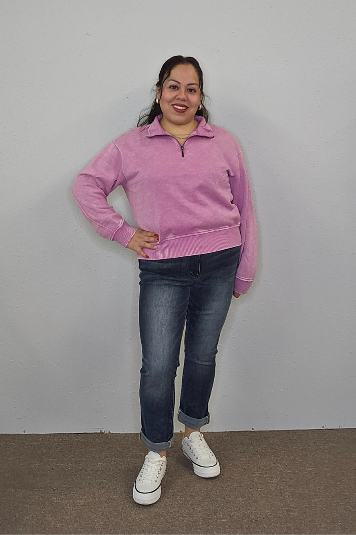 A woman stands smiling against a plain light wall, wearing the oversized cozy Acid Wash Half Zip in purple, rolled-up blue jeans, and white platform sneakers. One hand rests on her hip, and she wears bright red lipstick.