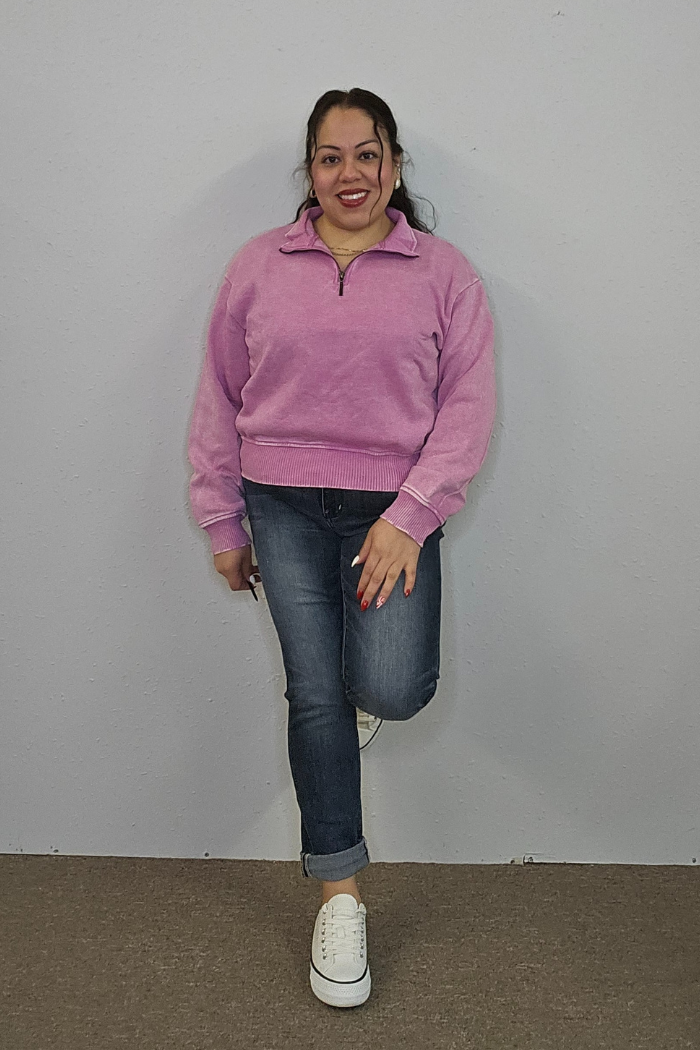A woman smiles against a gray wall, wearing the Acid Wash Half Zip in light purple with blue jeans and white sneakers. One leg is bent, her foot resting on the wall, and her hands are relaxed by her sides.