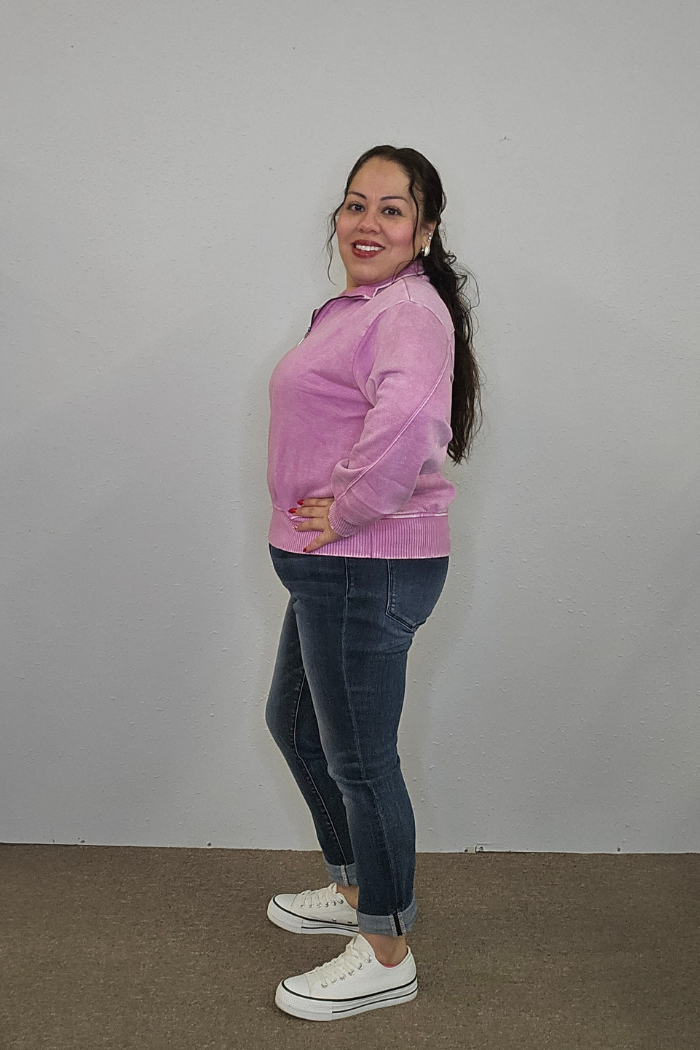 A woman stands sideways against a plain light wall, smiling. She is wearing the Acid Wash Half Zip pullover in pink, blue cuffed jeans, and white sneakers, with her hand on her hip and dark hair pulled back.