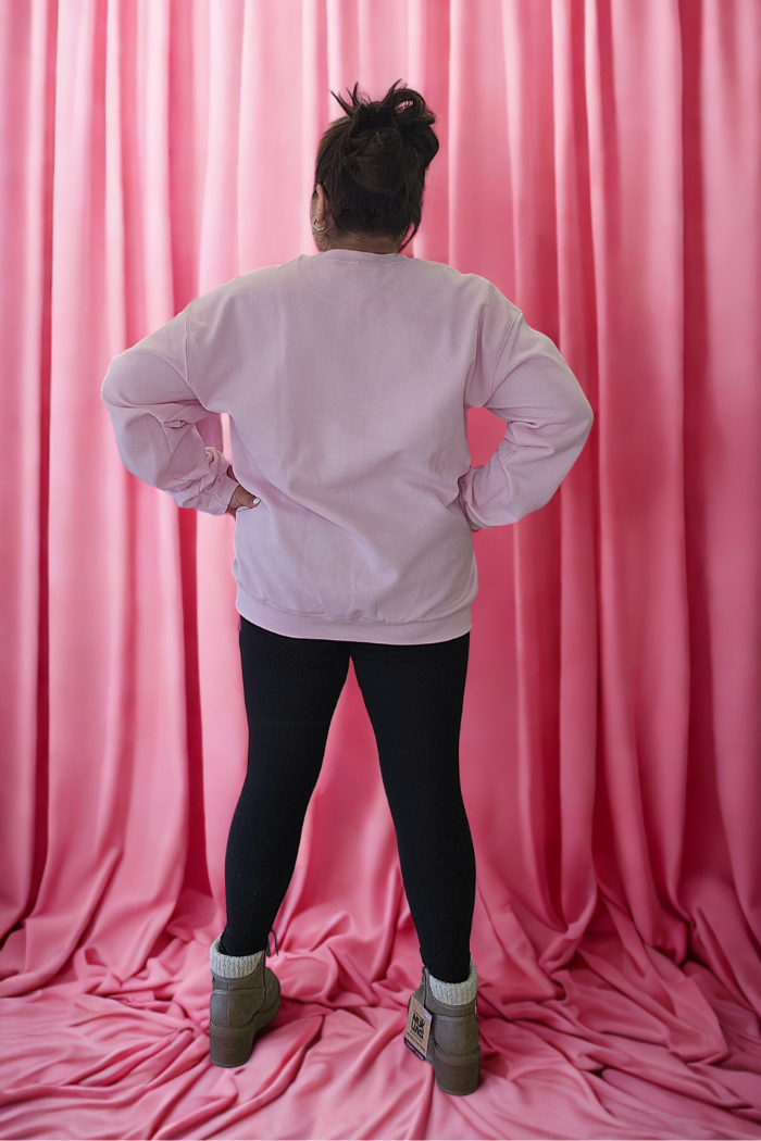 A person stands facing away with hands on hips, wearing the Love XOXO Crewneck in light pink, black leggings, beige boots, and gray socks. Soft pink fabric is draped from ceiling to floor in the background.