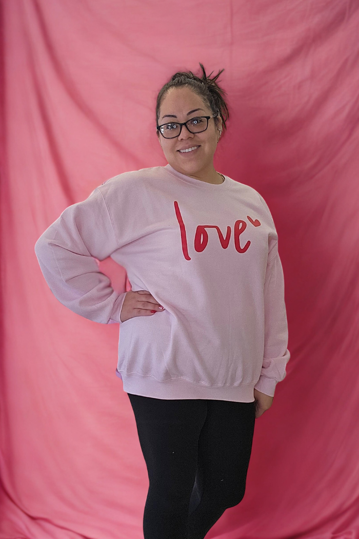 A woman stands in front of a pink background, smiling. She wears glasses, black leggings, and the Love XOXO Crewneck—a light pink oversized sweater with "love" and a small red heart—her hair tied up.