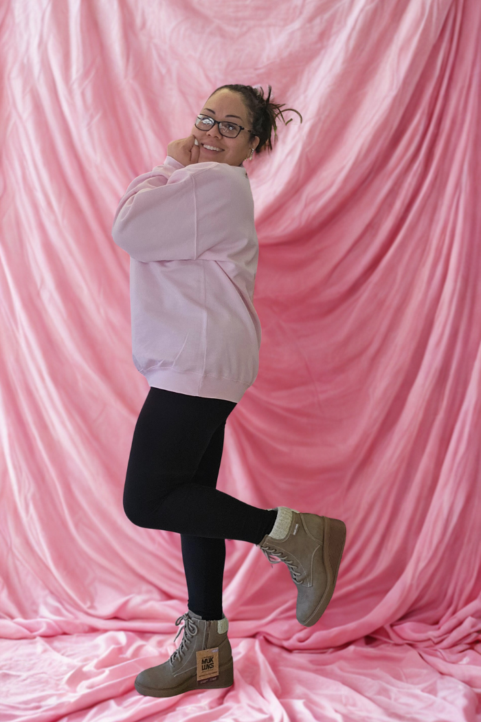A smiling person in glasses playfully poses on one leg in front of pink drapes, wearing the Love XOXO Crewneck sweatshirt, black leggings, tan boots, and cream socks.