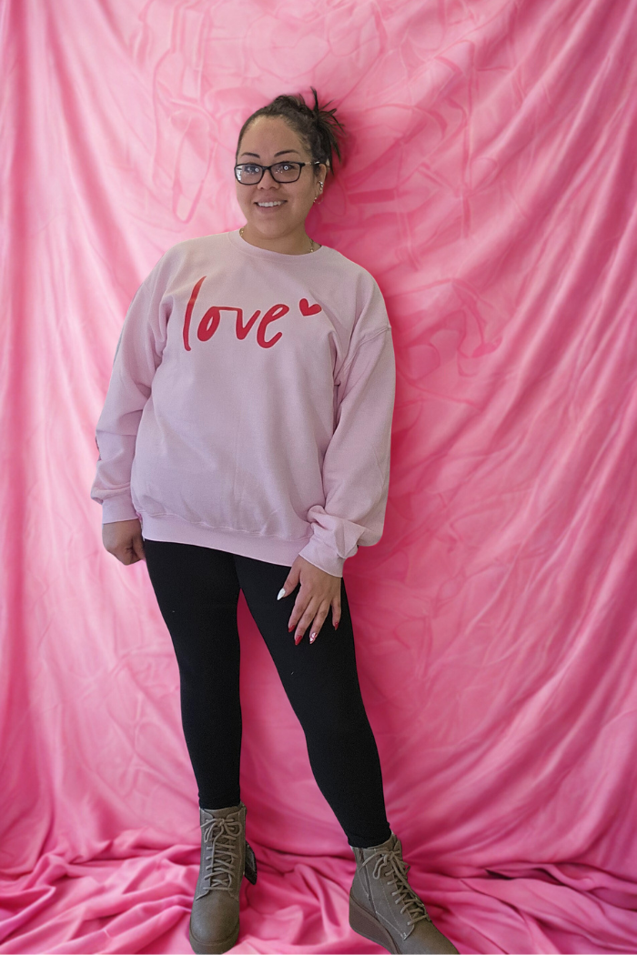 A woman smiles in front of a pink fabric backdrop, wearing the Love XOXO Crewneck with red “love” and heart lettering, paired with black leggings and tan boots. She wears glasses and has her hair pulled back for a cozy look.
