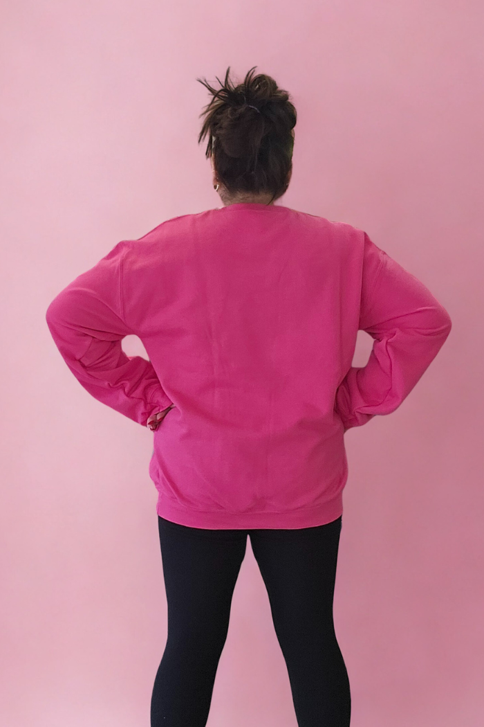 A person faces away from the camera against a pink background, wearing the bright pink Love XOXO Crewneck sweatshirt and black leggings with hands on hips.
