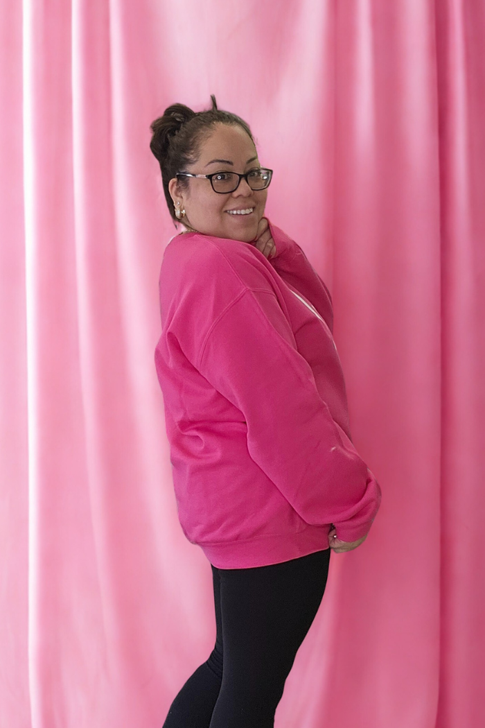 A woman wearing glasses and the bright pink Love XOXO Crewneck with black pants smiles while posing sideways in front of a matching pink curtain backdrop.