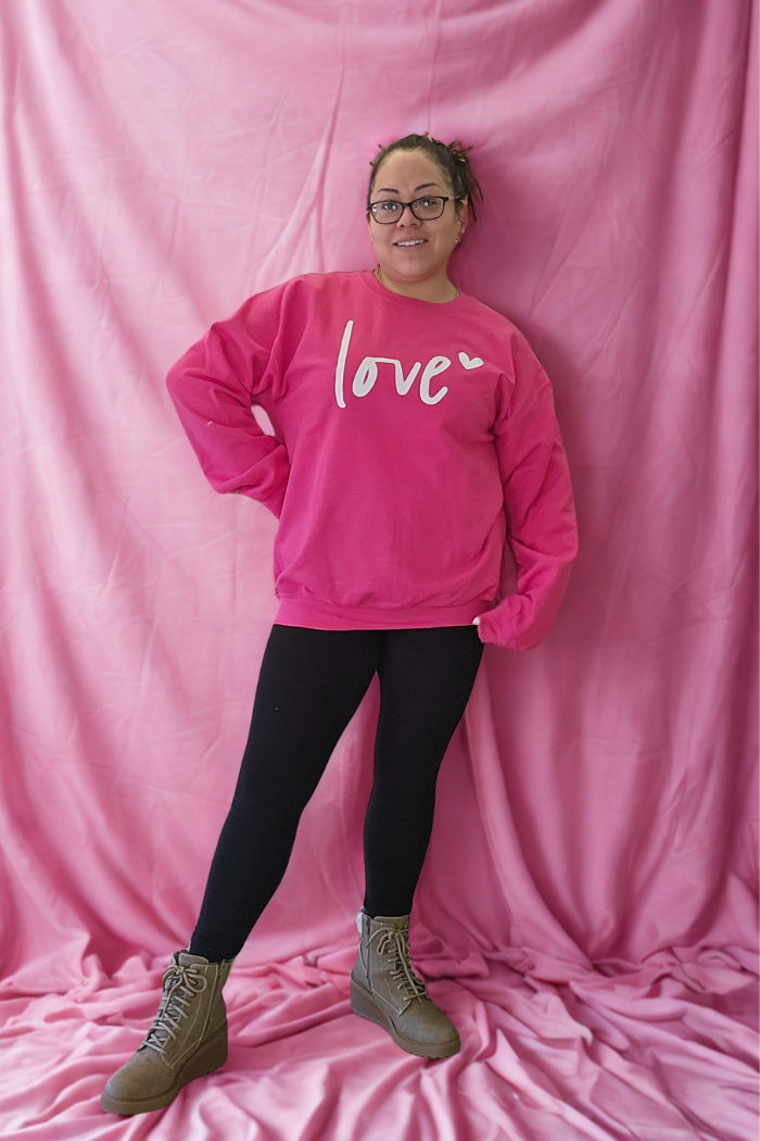 A woman stands on a pink backdrop wearing glasses, black leggings, gray boots, and the Love XOXO Crewneck—an oversized Valentine’s sweater with "love" on it—smiling with one hand on her hip.