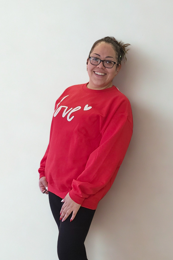 A smiling woman with glasses and a messy bun wears the Love XOXO Crewneck, featuring “Love” and a heart, paired with black leggings against a plain white background.