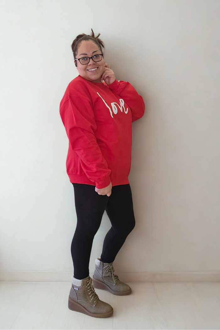 A smiling woman poses against a light wall, wearing glasses, black leggings, gray socks, brown lace-up boots, and an oversized Love XOXO Crewneck sweater.