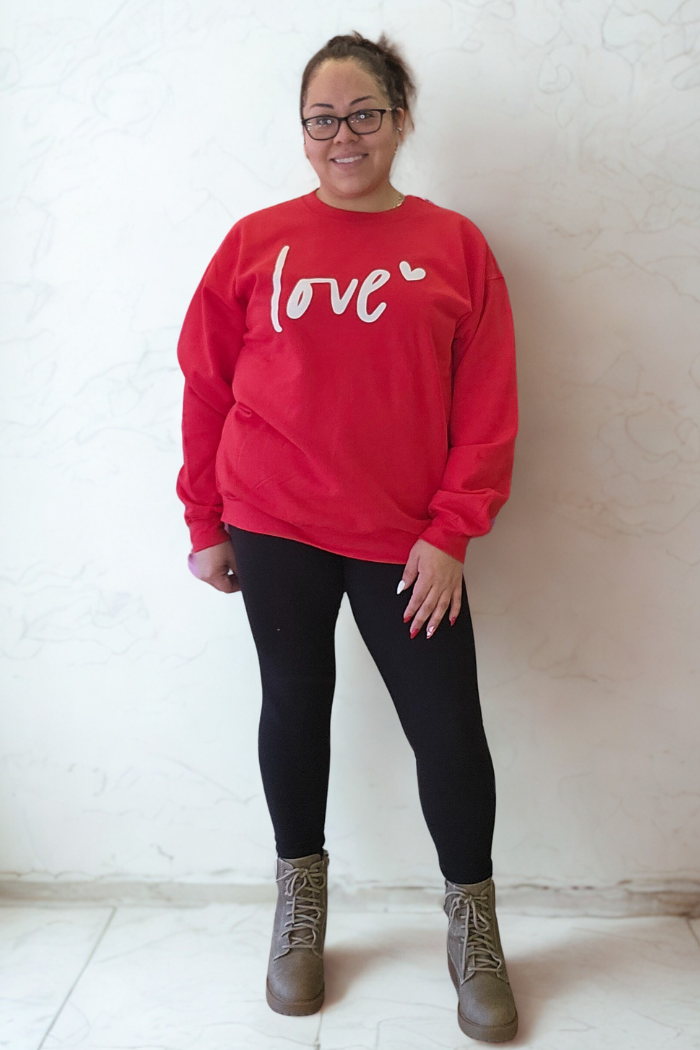 A woman wearing glasses, the Love XOXO Crewneck sweater, black leggings, and brown boots stands smiling against a light-colored marble-patterned wall.