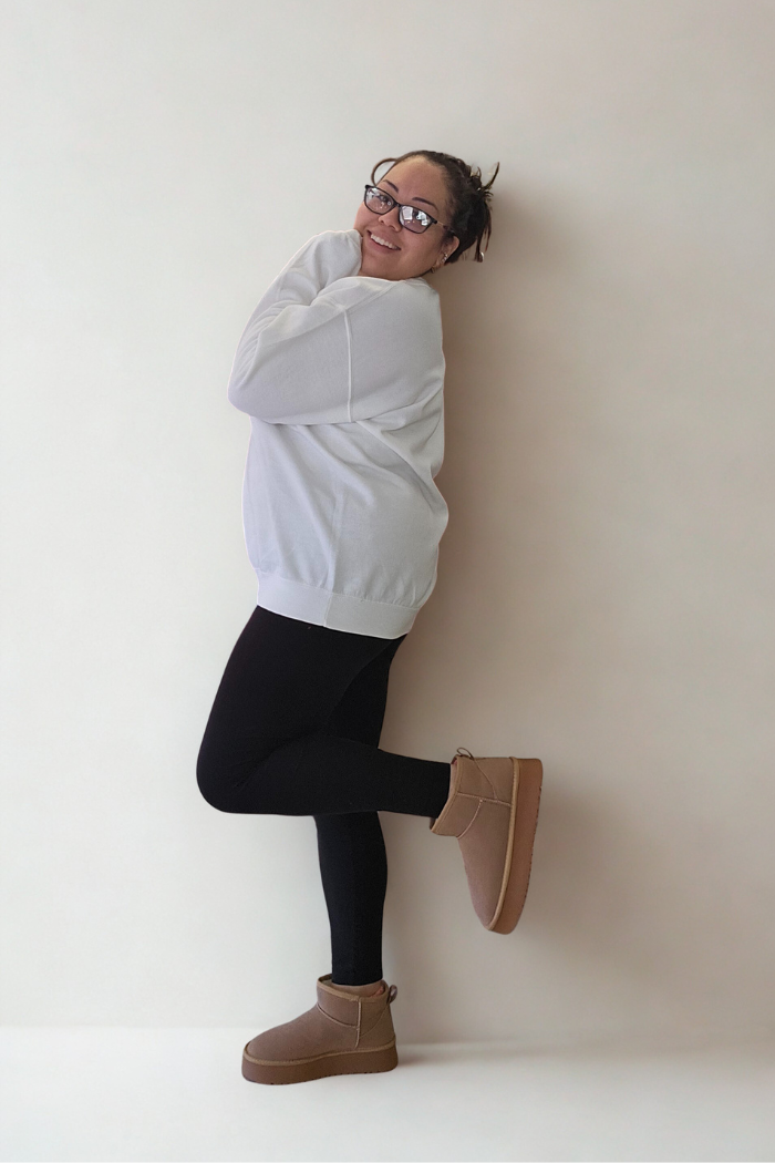A person in glasses, black leggings, tan boots, and a Lucky Mama Sweatshirt stands on one leg and smiles playfully against a plain light background.