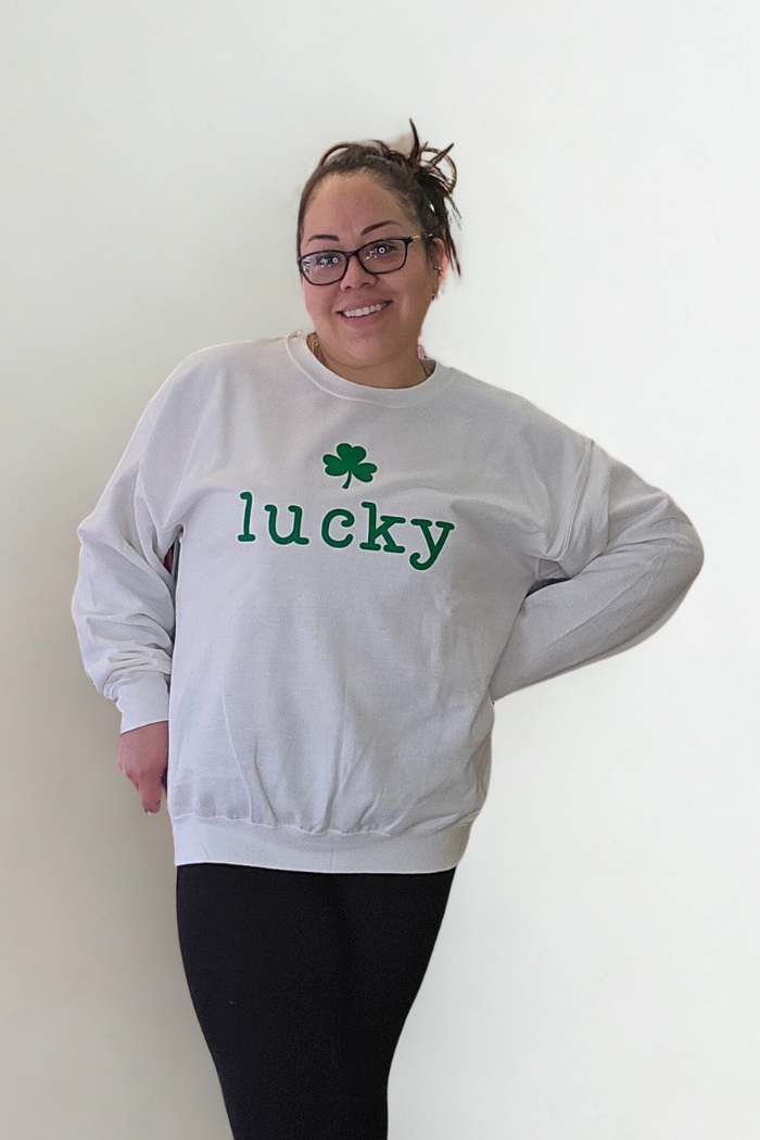 A smiling woman with glasses and a ponytail models the Lucky Mama Sweatshirt featuring a green shamrock and "lucky" print, paired with black leggings—ideal St. Patrick’s Day attire—against a plain white background.