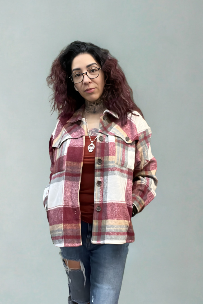 A person with wavy dark hair, glasses, and tattoos wears the Plaid Button-Up Shacket in red and beige over a rust-colored top and ripped jeans against a plain light gray background.