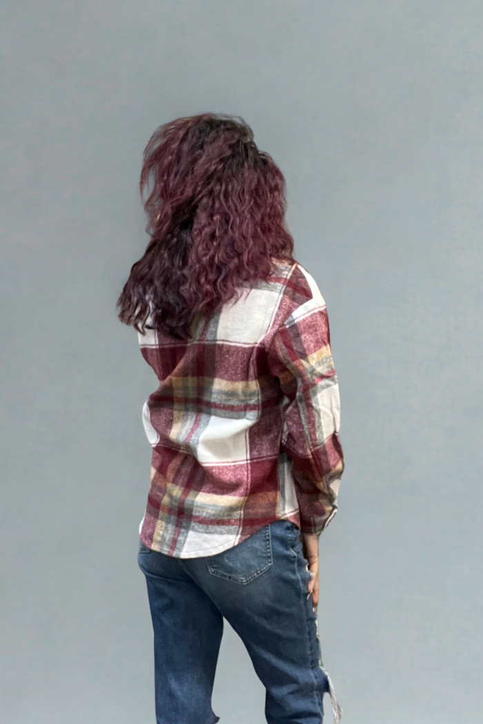 A person with wavy, dark hair faces away, wearing a red and white Plaid Button-Up Shacket and blue jeans against a plain gray background.