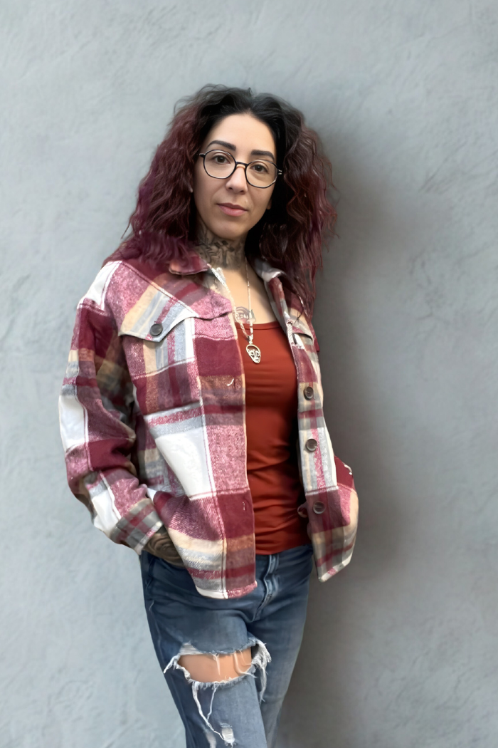 A person with long, curly dark hair and glasses stands against a gray wall, wearing the Plaid Button-Up Shacket over a rust-colored top, paired with ripped jeans and necklaces, hands in jacket pockets.