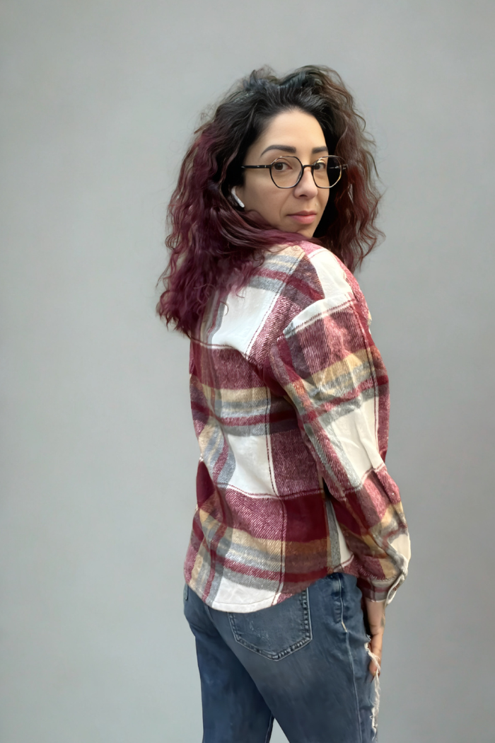 A person with wavy dark hair and glasses wears a red Plaid Button-Up Shacket, standing slightly turned to the side and looking over their shoulder against a plain gray background.