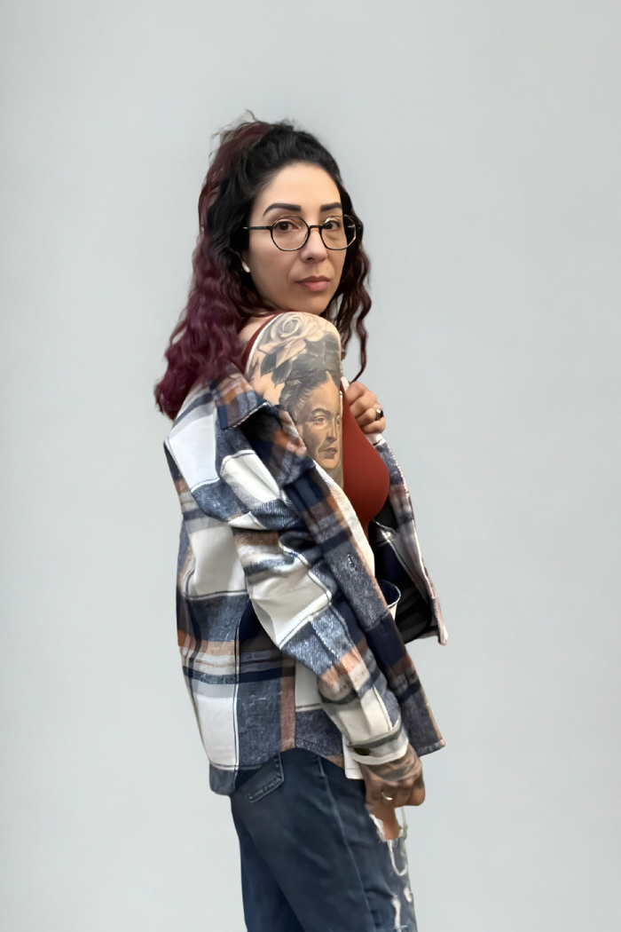 A woman with dark wavy hair and glasses models the Plaid Button-Up Shacket over a brown top, standing sideways to show a large tattoo of a woman's face on her upper arm against a plain light gray background.