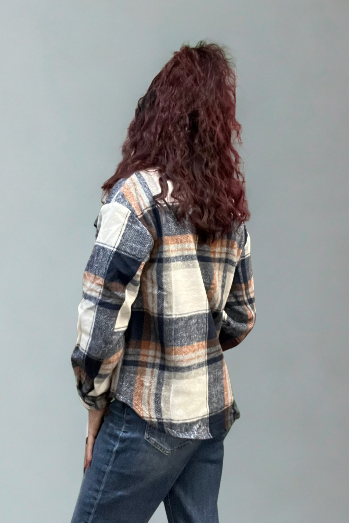 A person with long, curly, reddish-brown hair faces away, wearing the Plaid Button-Up Shacket in blue, beige, and orange with blue jeans against a plain gray background.