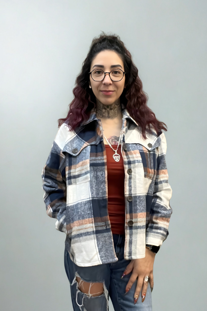 A young woman with long, wavy dark hair and glasses stands against a plain background, wearing a Plaid Button-Up Shacket over a brown top with ripped jeans; tattoos are visible on her neck and hands.