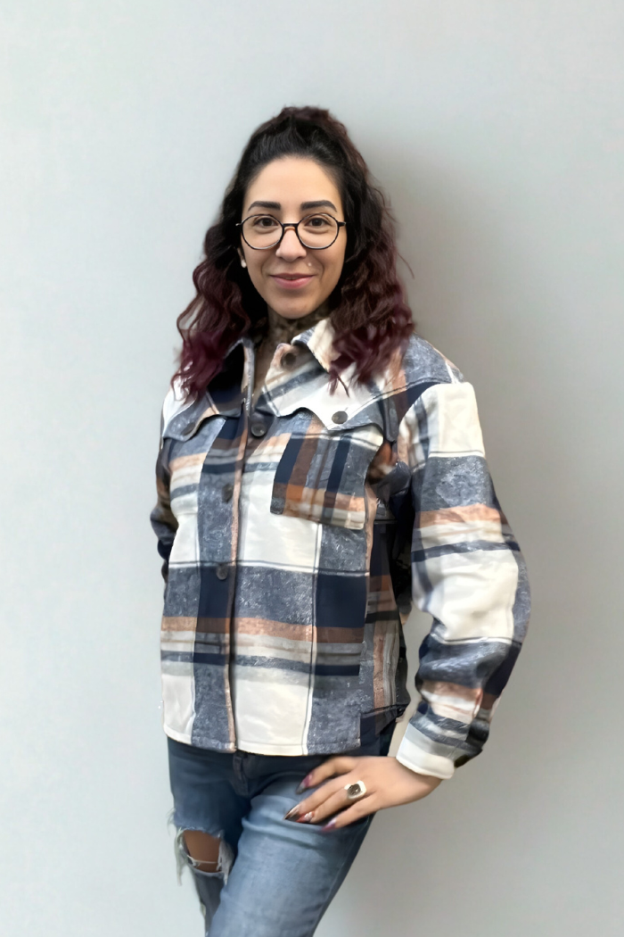 A woman with curly dark hair and glasses smiles at the camera, wearing a Plaid Button-Up Shacket over ripped jeans, standing against a plain light gray background.